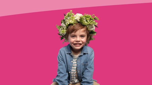 Bambino con corona di fiori in testa e giacca di jeans seduto su sfondo rosa acceso.