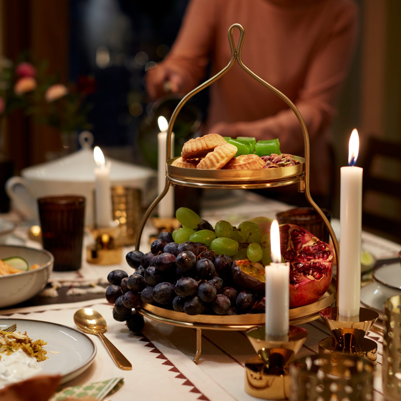 حامل تقديم، دورين GOKVÄLLÅ بلون ذهبي يحمل عنب ورمان وبسكويت، على طاولة مزينة بالشموع.