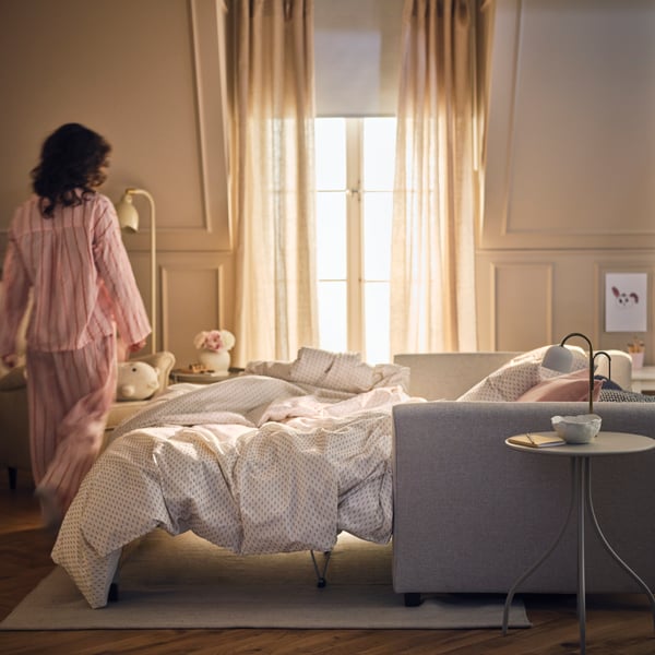 Una persona junto a un sofá cama de 2 plazas EKHOLMA en beige claro configurado como cama, cerca de una ventana con cortinas DYTÅG.