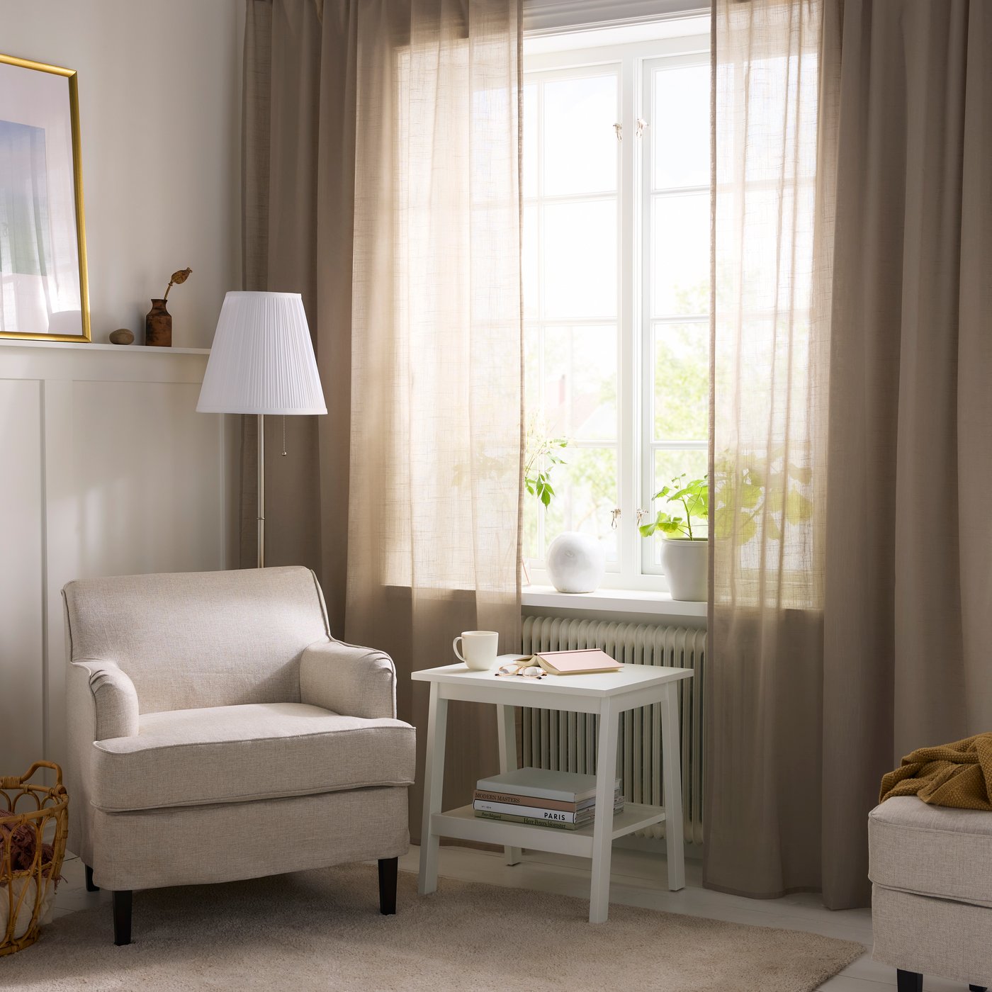 Ett ljusbeige vardagsrum med tunna beige gardiner i dst stora fönstret sdär vårljuset silas in. 