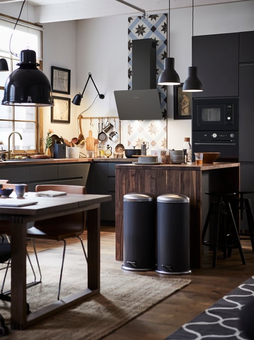 Kitchen setting with a grey base cabinet with three drawers, pedal bins, a table with a brown chair, and a work wall lamp.
