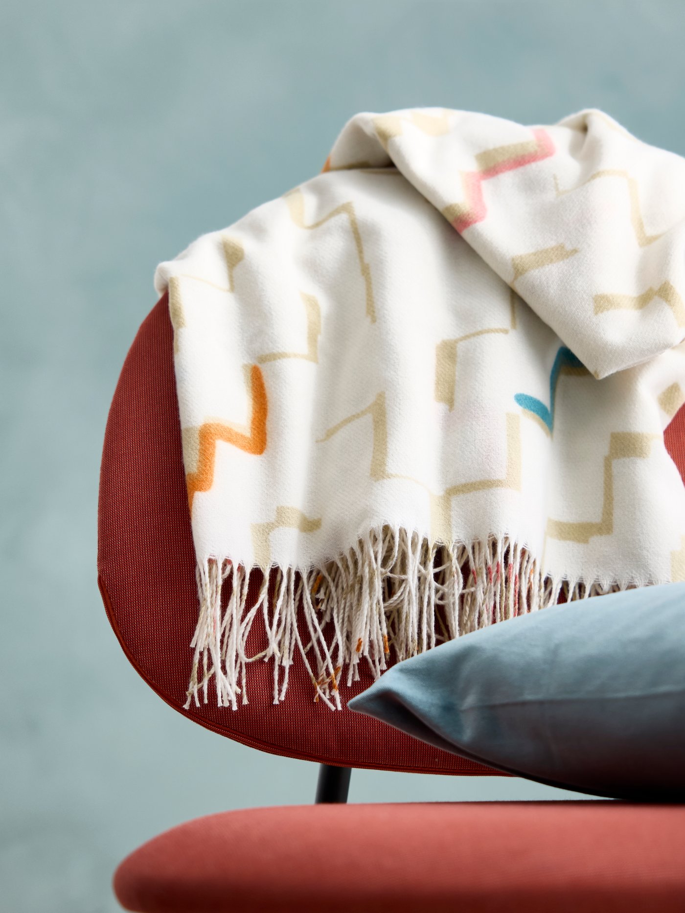 Close up of a OLIVSPINNMAL throw and a SANELA pillow placed on a red GÄVLE armchair.