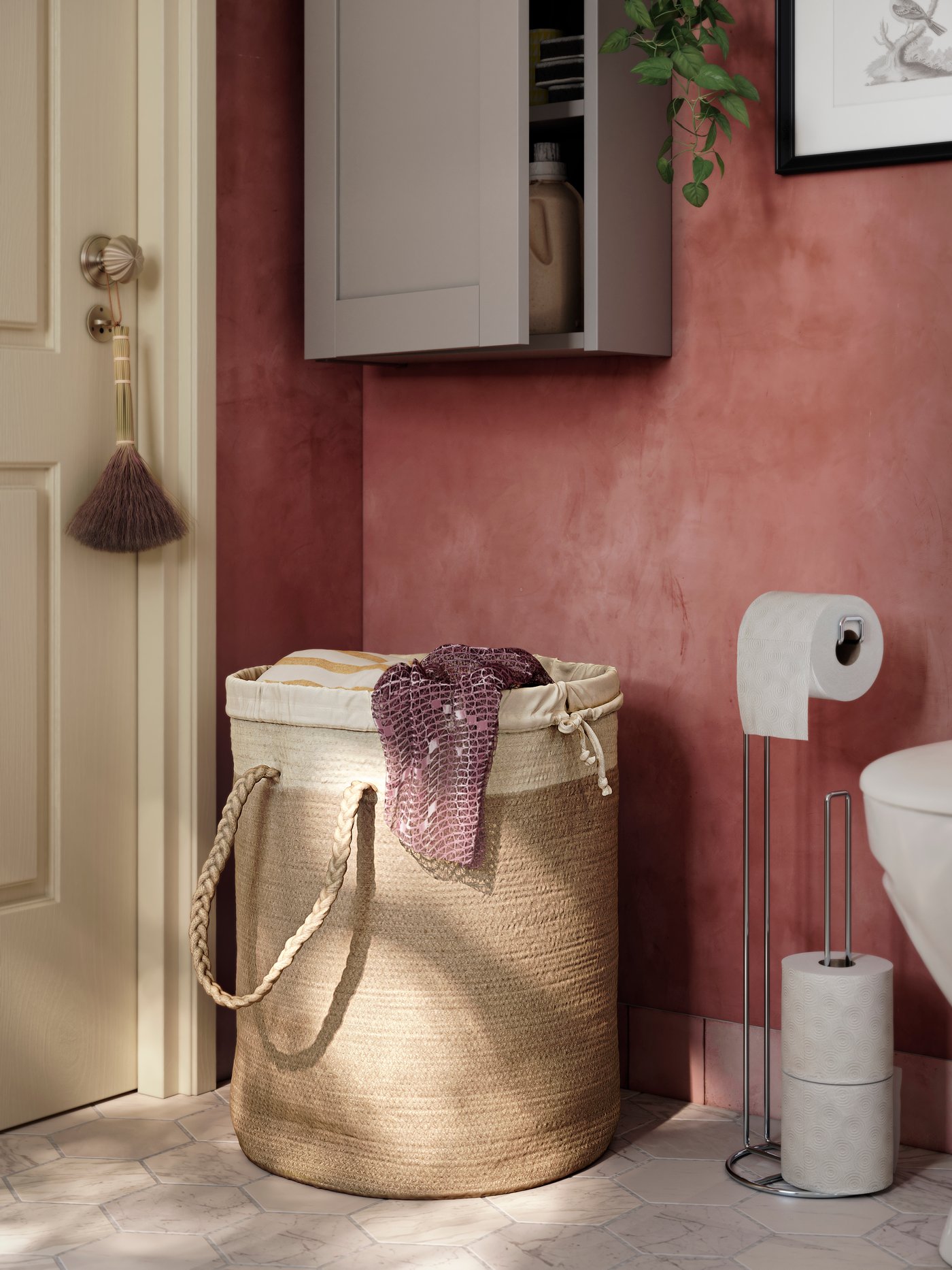 Standing in a corner of a pink bathroom, underneath a grey wall cabinet, is a LJUNGAN laundry basket in natural materials.