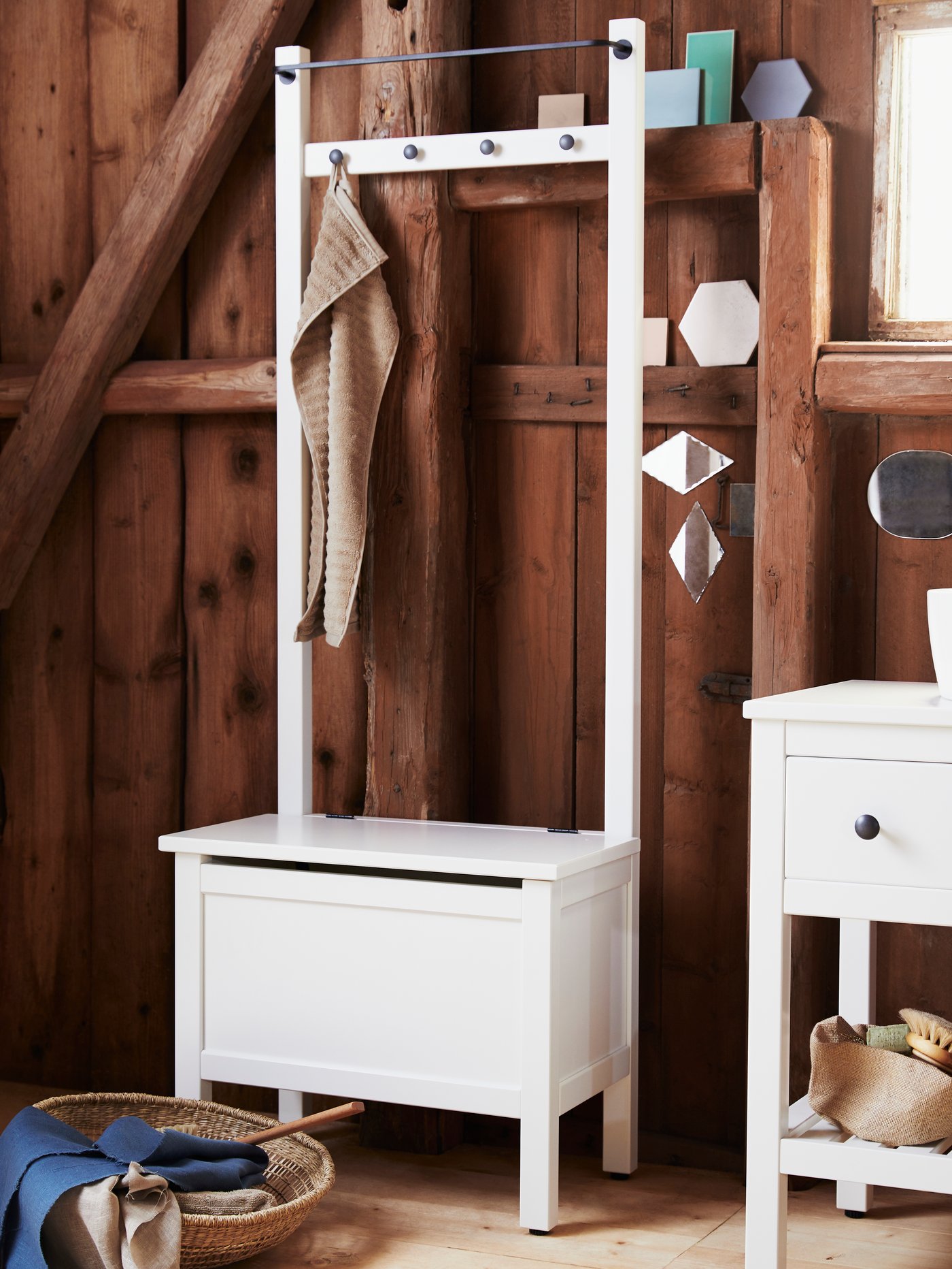 A white HEMNES storage bench, a towel hanging from a hook, a basket with blue linen and beige towels on a wooden floor.