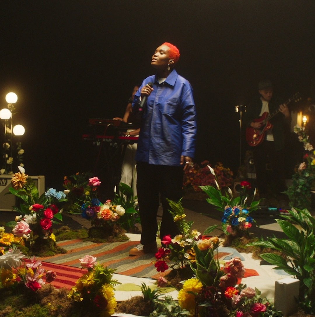 Muzičarka Arlo Parks s mikrofonom, okružena cvećem.