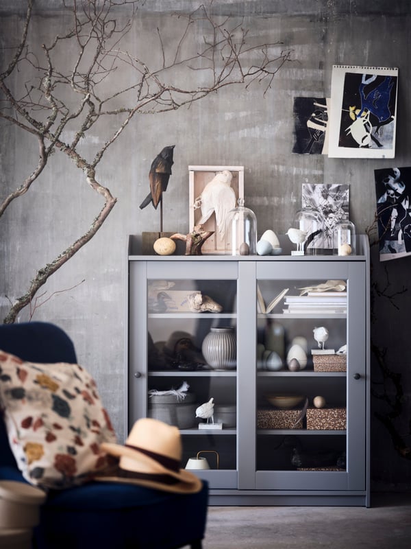 A grey HAUGA glass-door cabinet is filled with bird-themed decorative items, boxes and glass domes, in a grey living room.