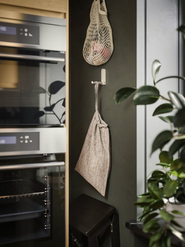 A KLYKET folding hook in aluminium and beige, with a bag hanging from it, is attached to a kitchen wall next to a window.