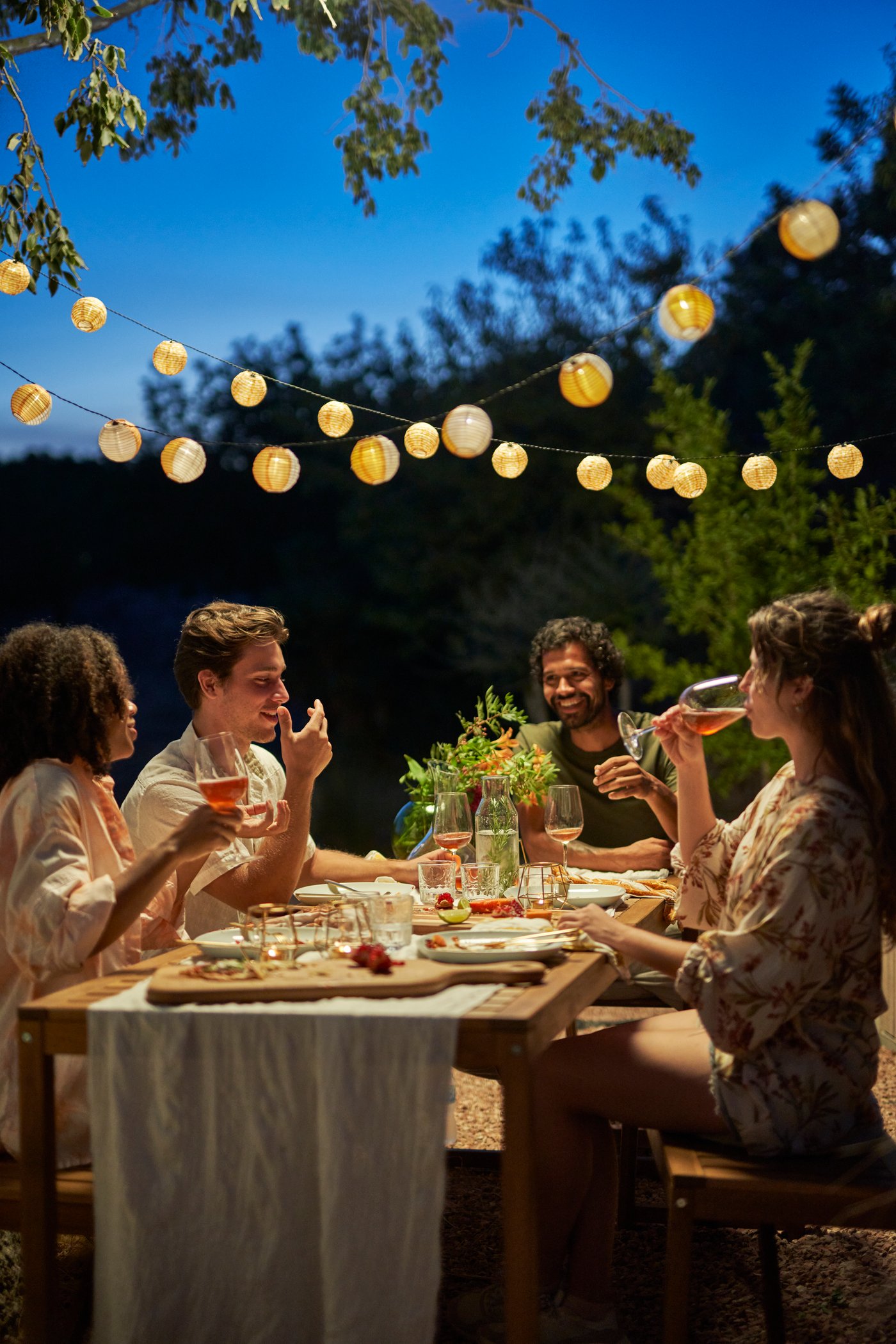 En gruppe sidder og nyder et aftensmåltid udendørs under en SOMMARLÅNKE LED lampe.