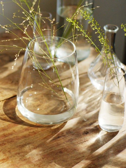 A wooden table holds a group of mixed clear glass vases, including a curved BERÄKNA clear glass vase with a plant sprig.