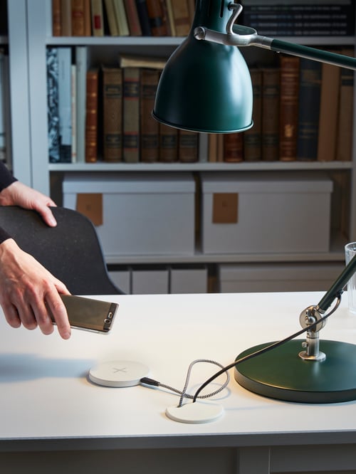 Un prim-plan al unei persoane în spațiul de lucru cu un încărcător wireless alb subțire proiectat pe un birou dreptunghiular și o bibliotecă albă.