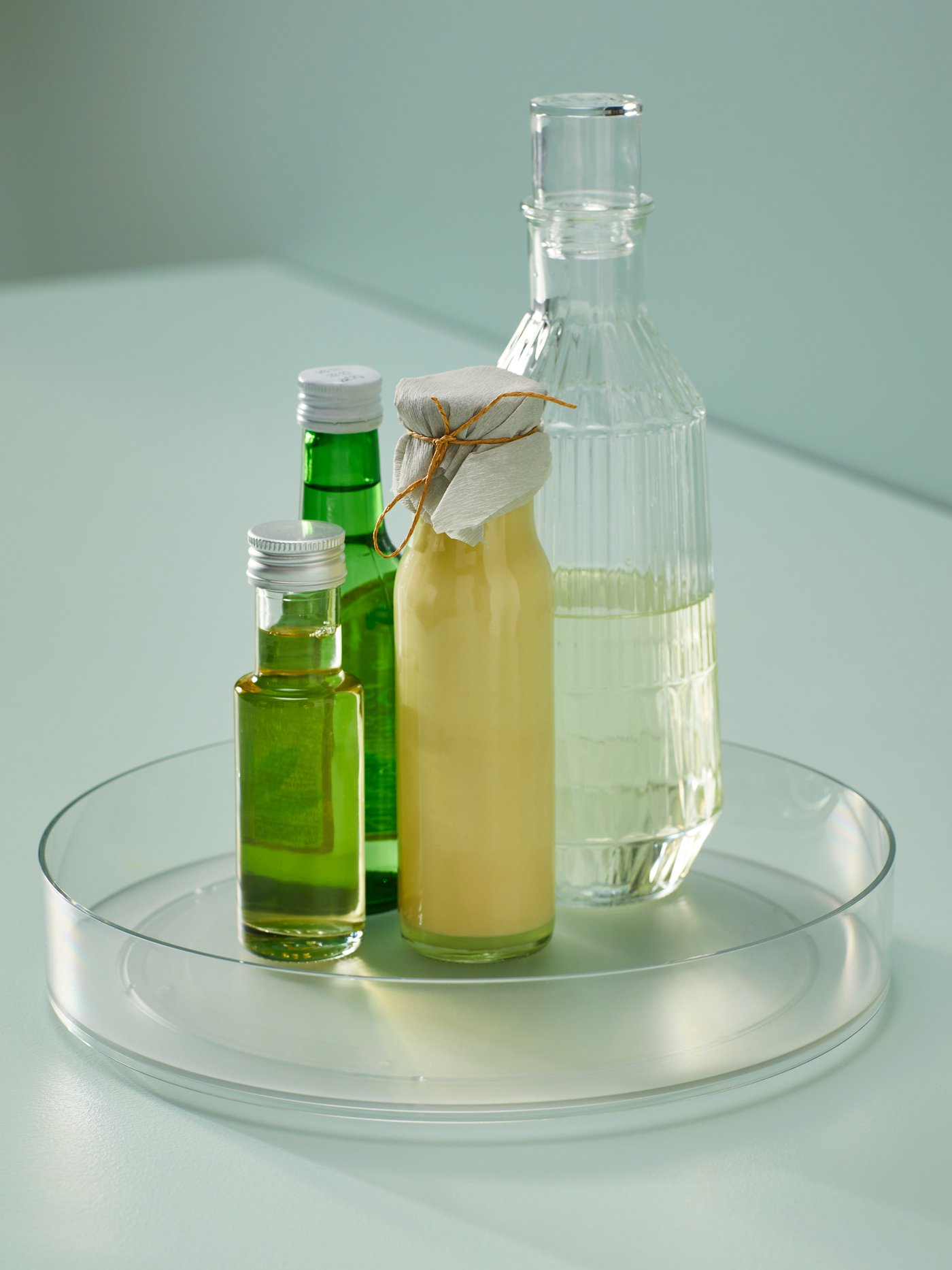 Na prozirnoj plitkoj posudi stoje tri staklene boce: dvije zelene manje boce, jedna veća prozirna boca te žuta boca s papirom i vezicom na čepu.