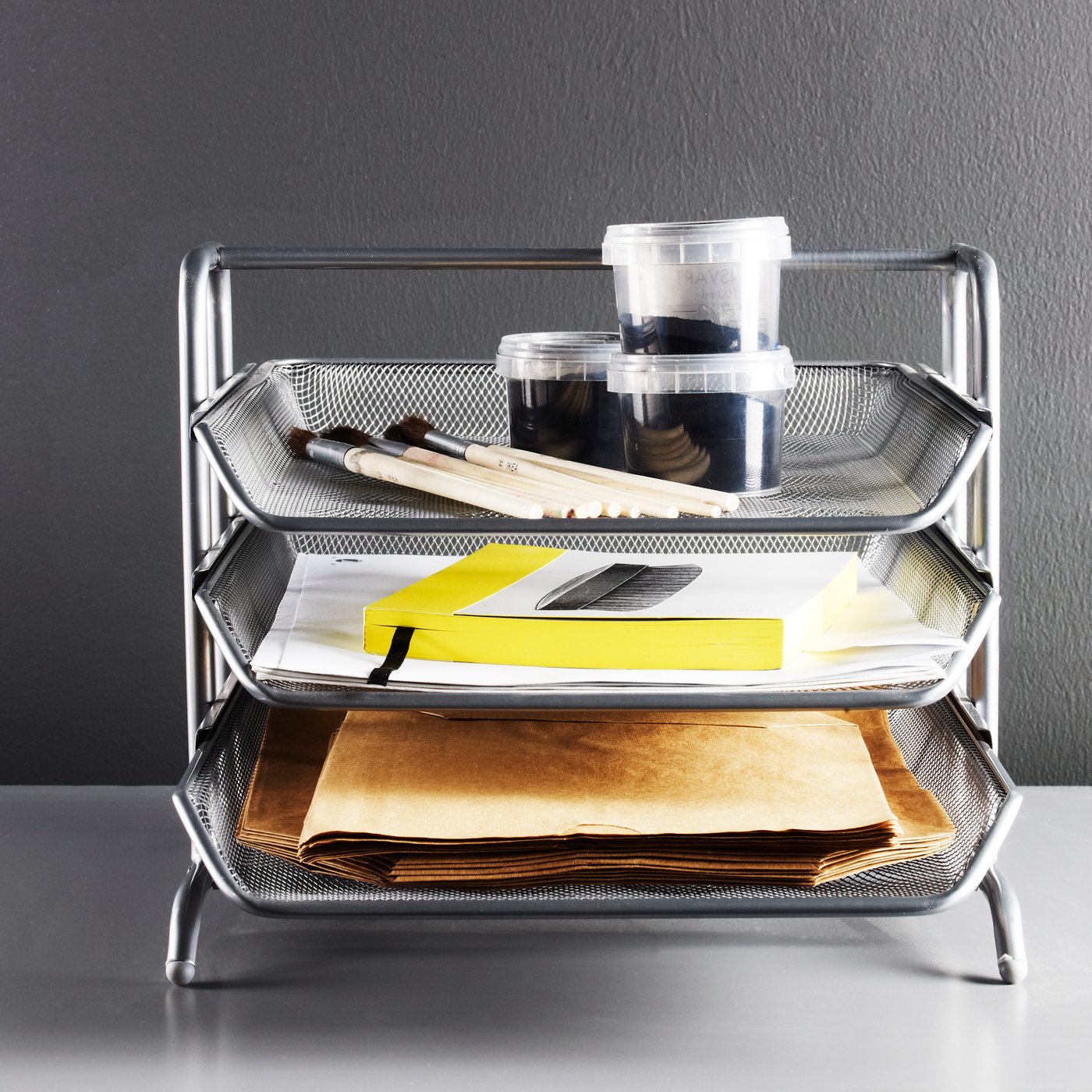 A metallic letter tray with three levels, holding plastic jars, files and various items placed on a white desk.