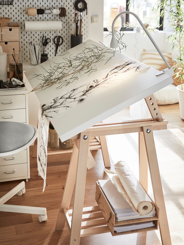 Valkoinen kallistettu pöytälevy puunväristen jalkojen päällä. Pöydässä on kiinnitettynä valaisin ja sen päällä on käsityö. 