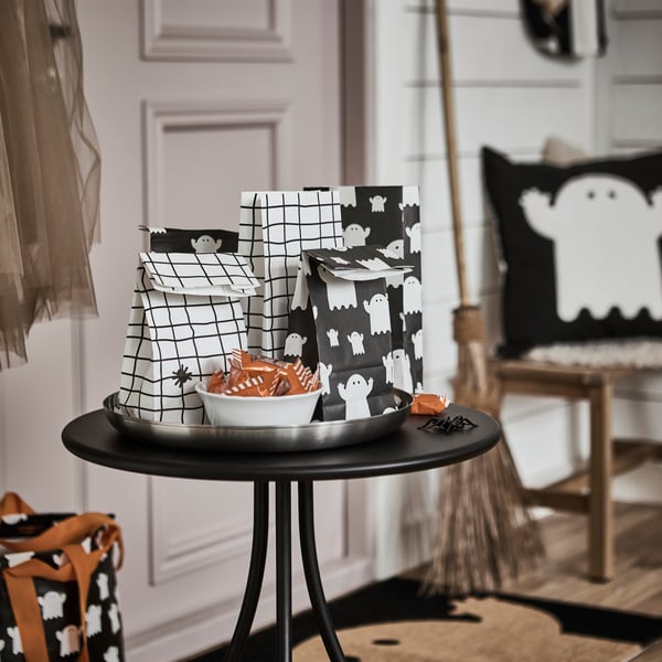 Some KUSTFYR paper bags featuring ghosts and spiders lie on a small black table in a hallway, next to a bowl full of treats.