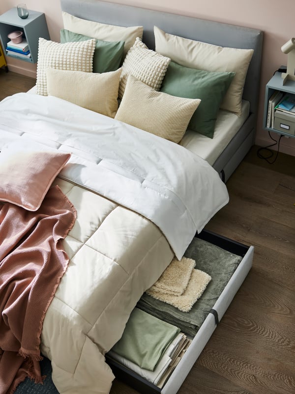 A grey GLADSTAD upholstered bed with a mix of cream, green and pink bed textiles on it including a HEDFRYLE comforter.