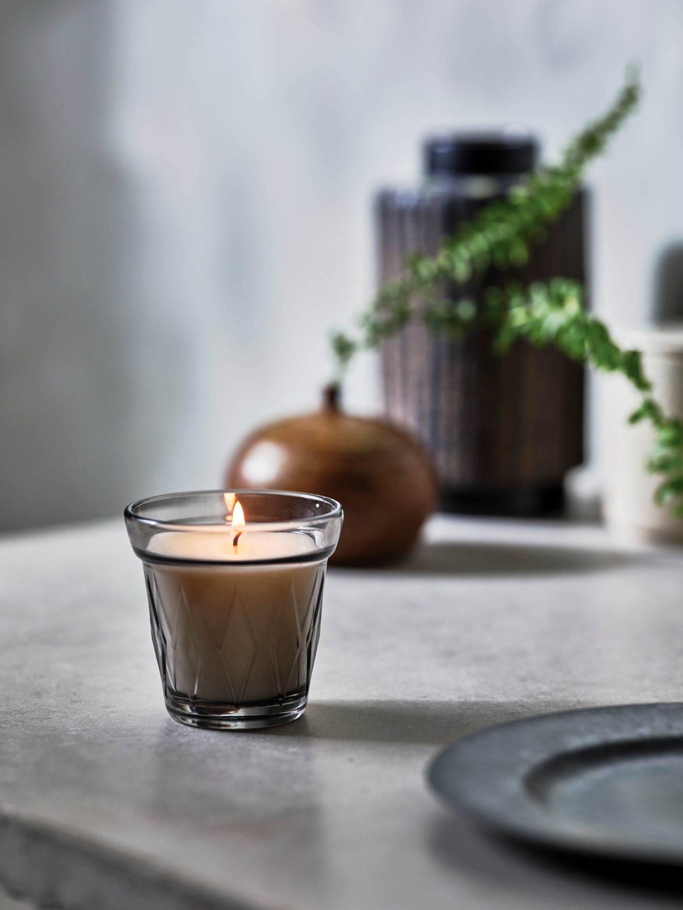 A lit VÄLDOFT scented candle in glass stands near a plate and a vase with a small plant in it and another vase.