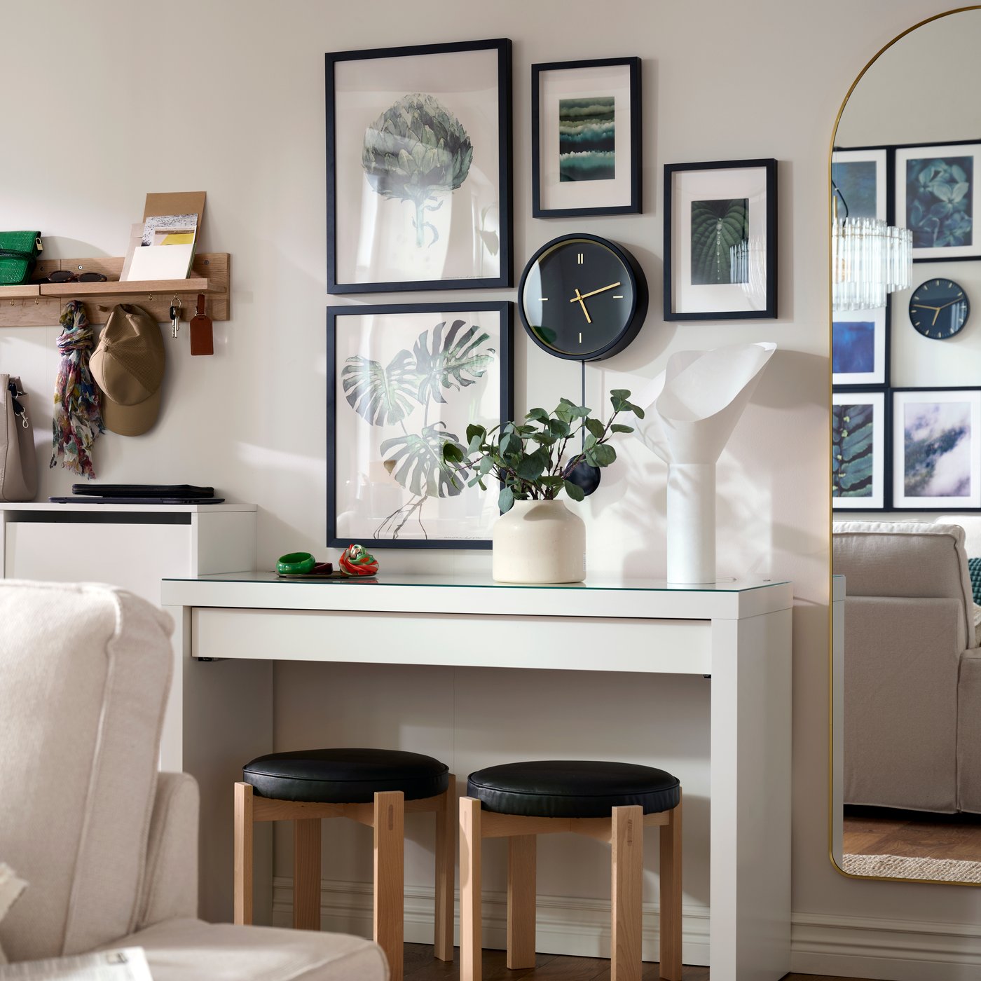 Framed art hanging around a black STURSK wall clock, all placed above a MALM dressing table with a BÄCKNATE table lamp on it.