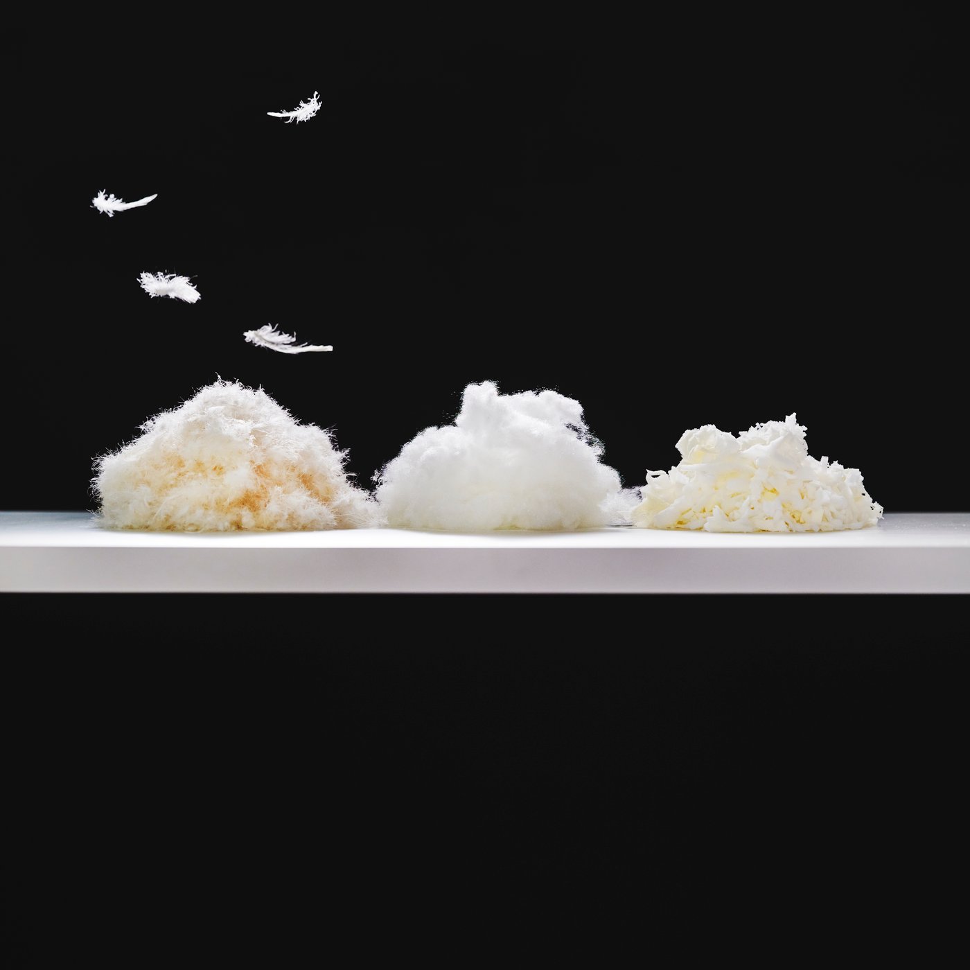 Three small piles of down and feathers, polyester pillow filling and shredded memory foam side by side on a white shelf.
