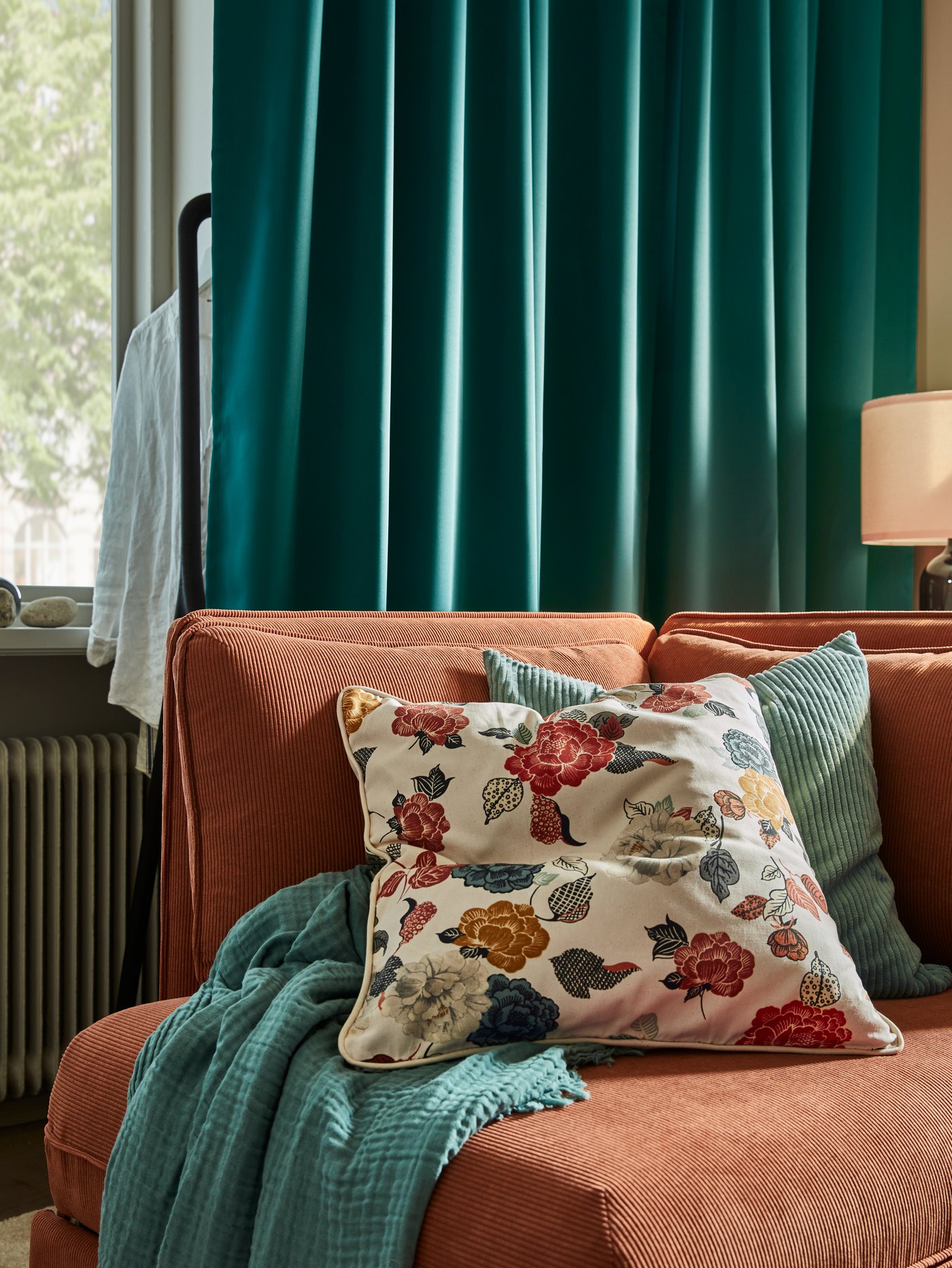An orange sofa-bed with a floral patterned cushion, a teal corduroy cushion, a teal throw and blue-green curtains.