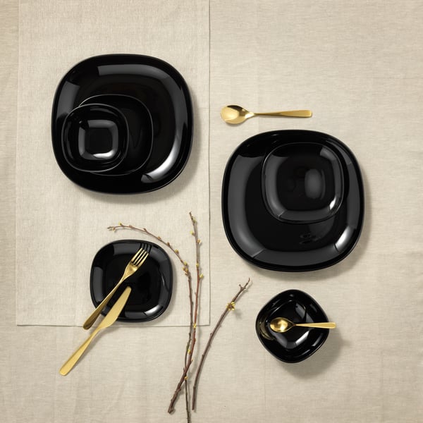 Four black IKEA BACKIG plates of various sizes on top of a beige table cloth, gold cutlery across.