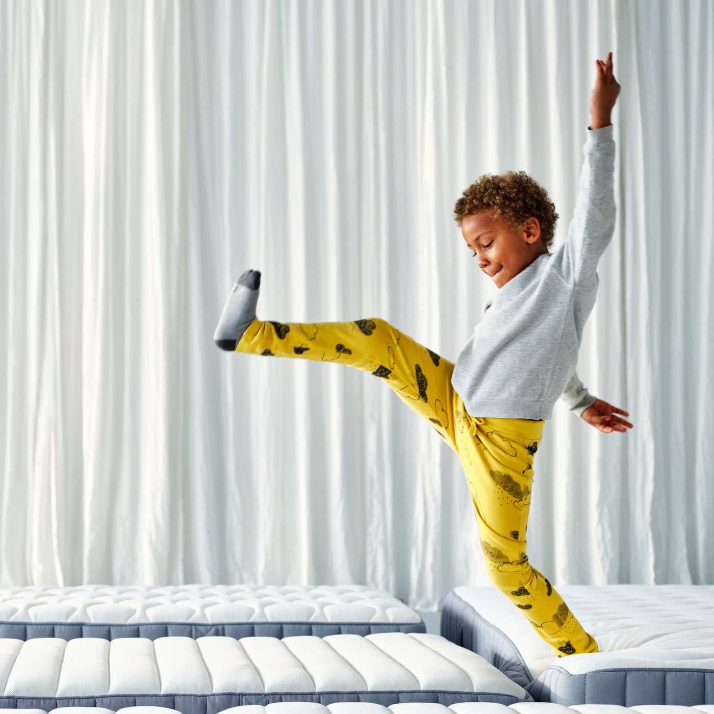 A young boy in bright yellow pyjamas trousers and a white top is jumping from one mattress to another in a white studio space.