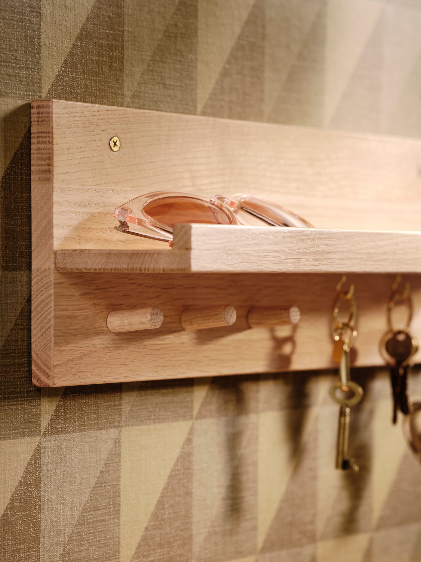 Un organizator HÄSTVISKARE din lemn masiv de stejar pe un perete cu model, cu brelocuri de chei agățate de cârlige și o pereche de ochelari de soare pe poliță.