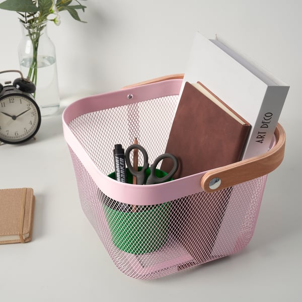 Ein rosa RISATORP-Korb gefüllt mit einem Buch einem Notizbuch und einem Behälter mit Stift und Scheere, außerhalb ein Wecker mit einer kleinen Vase und Blumen