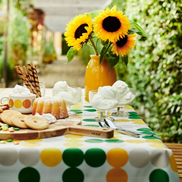 Table avec nappe à pois BRÖGGAN sur laquelle sont disposés de la vaisselle, des bonbons, des biscuits, des desserts et un vase jaune garni de tournesols.