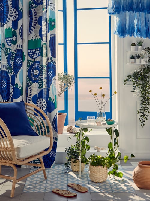 A BUSKBO armchair in rattan material, next to a tray table and multicolour curtains, in front of an open window.