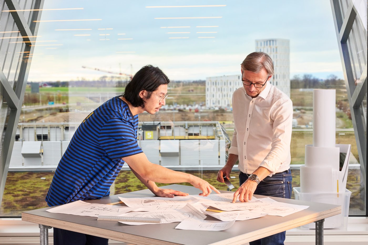 Dva muškarca pregledavaju nacrte na stolu u modernom uredu s velikim prozorima i pogledom na gradilište.