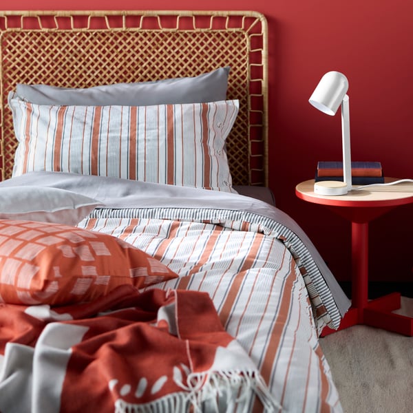 Une lampe de bureau posée sur une table d’appoint rouge, à côté d’un lit avec une tête de lit en rotin, habillé de la parure de lit GULDAKLEJA rouge brun.