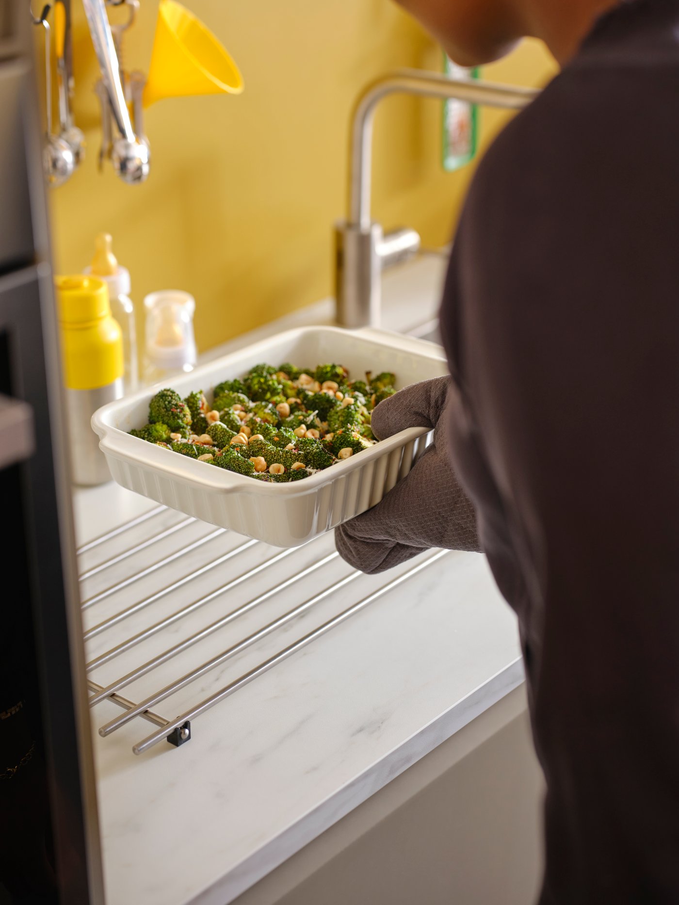 A hand with an oven glove puts an off-white oven dish on a LÄMPLIG stainless steel trivet on a white marble effect worktop.