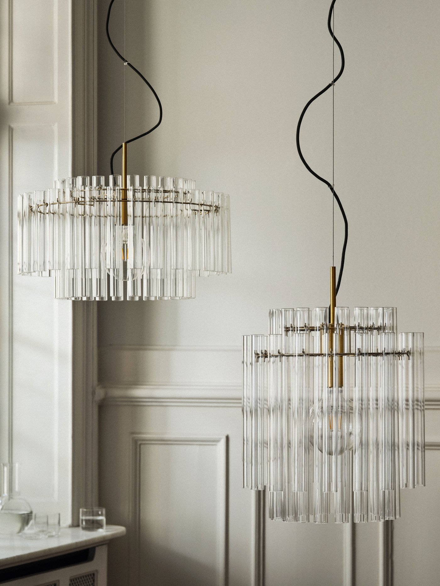 Två STOCKHOLM 2025 taklampor i glas med mässingsplätering hänger bredvid varandra i ett beiget rum.