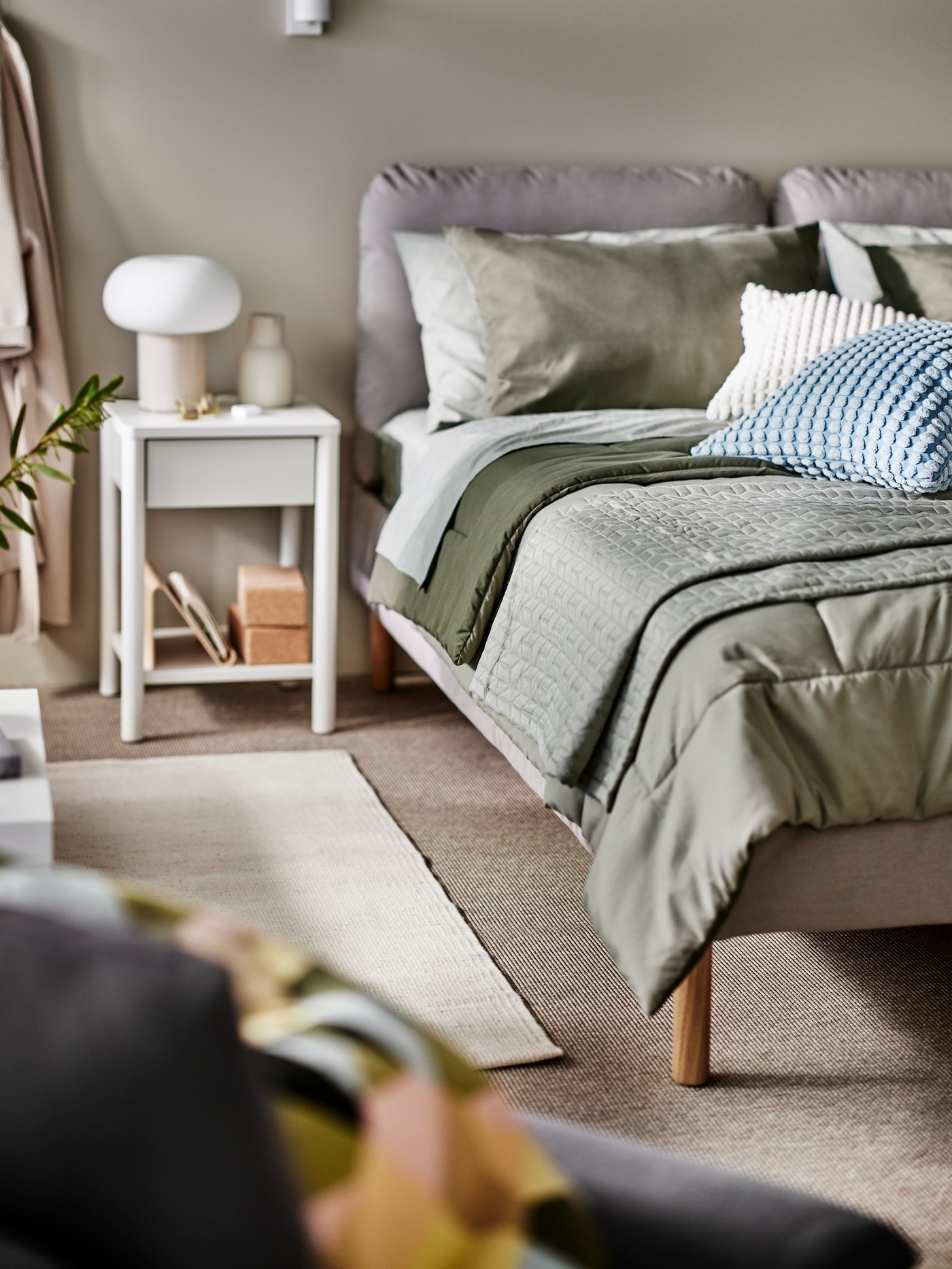 An upholstered bed with a green HEDFRYLE 3 comforter and other bed linen on it and an off-white TONSTAD bedside table by it.