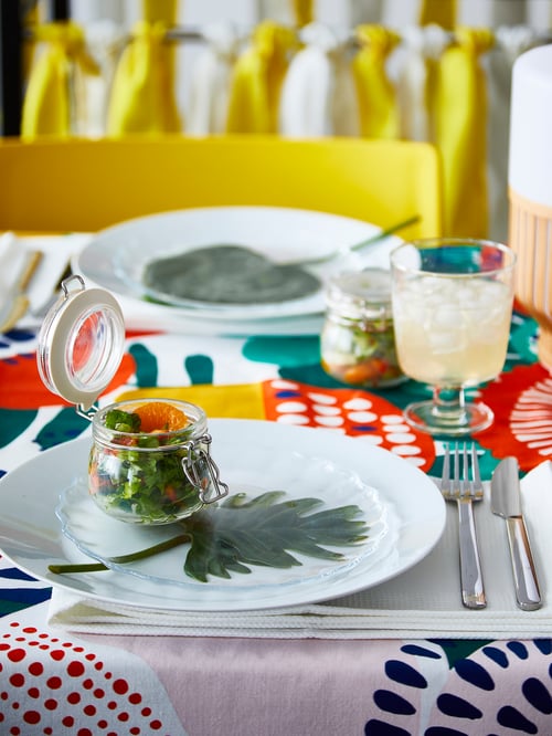 Un couvert est disposé sur une nappe aux motifs vibrants avec un verre plein et un bocal avec couvercle KORKEN en verre transparent empli de salade.