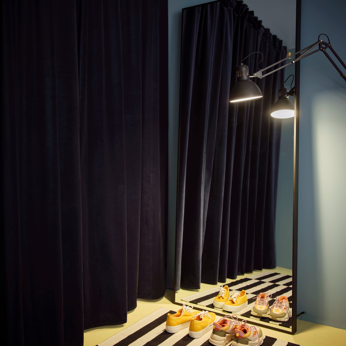 HOVET mirror leans against a wall, reflecting dark curtains, a floor lamp and colourful shoes on a striped rug.