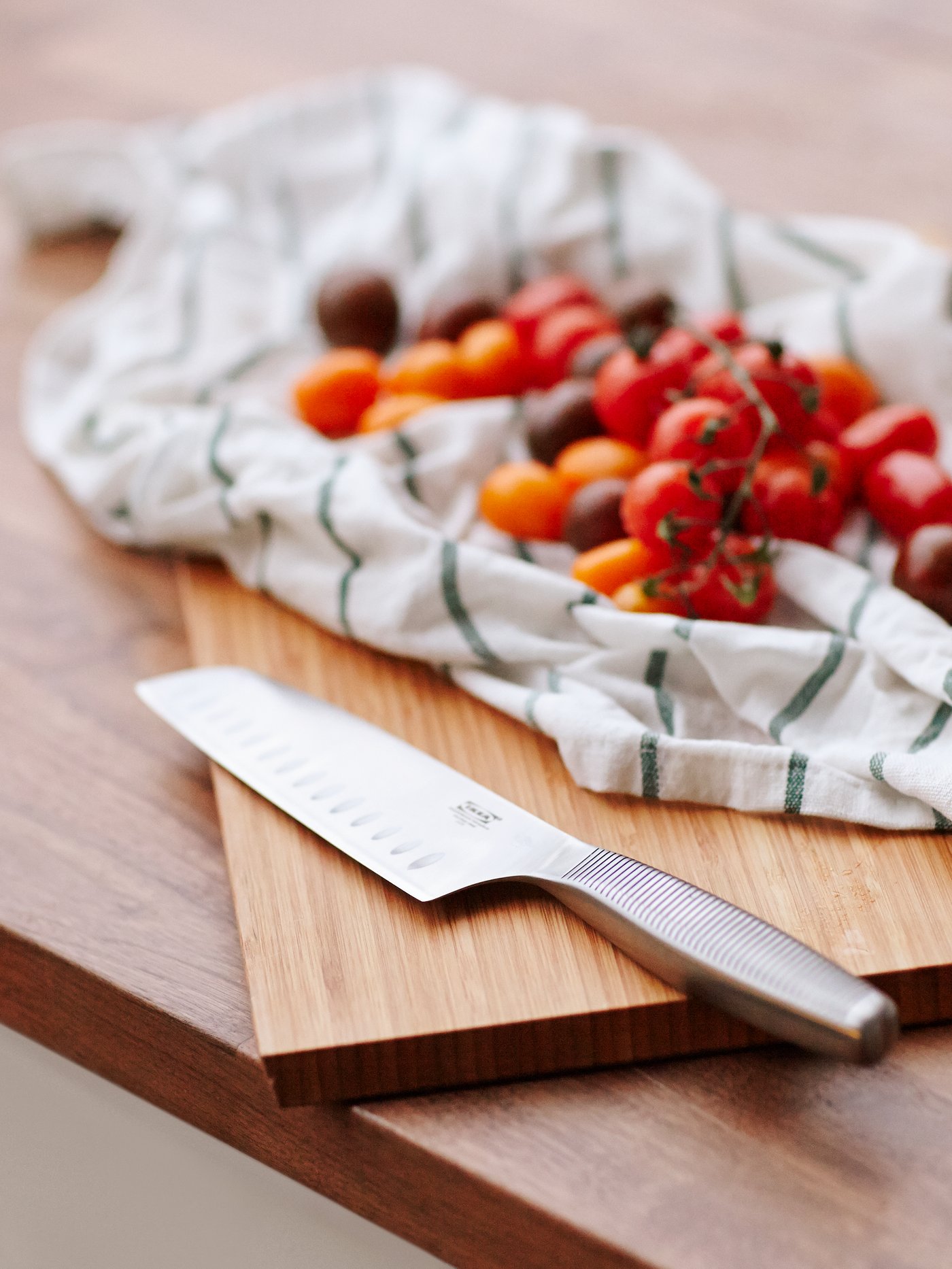 Close-up of a grey vegetable knife on a beige chopping board, set next to cherry tomatoes on a tea towel.