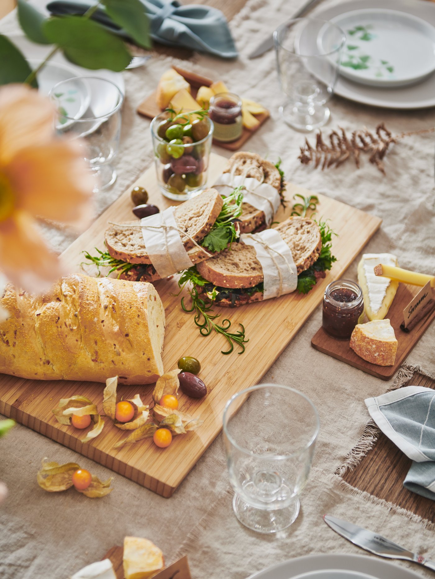 Mesa con una tabla de cortar beige, paños de cocina planos, platos de porcelana blanca con estampado verde y cubiertos.