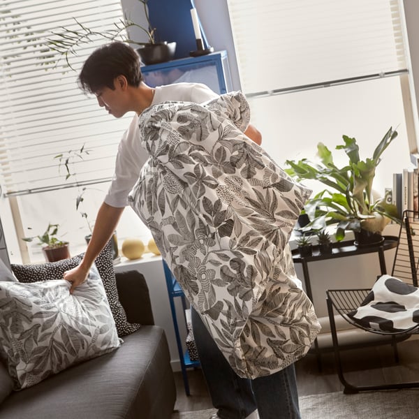 A teenager holding a BROKDRACENA duvet cover with a black and white leaf pattern while placing a pillow on a sofa-bed.