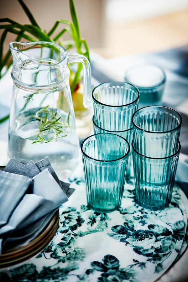 Bord dækket til frokost, fyldt med glas, brød, servietter, tallerkener, en grøn bakke og et skærebræt af træ.