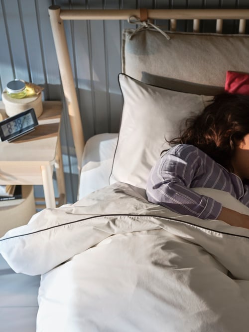 Bedroom close-up of a person laying on a beige bed frame made of birch, covered by a duvet cover with 2 pillowcases.