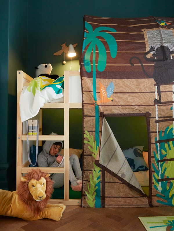 A child sits with a book inside a KURA reversible bed with a KURA bed tent with curtain, next to a DJUNGELSKOG lion.