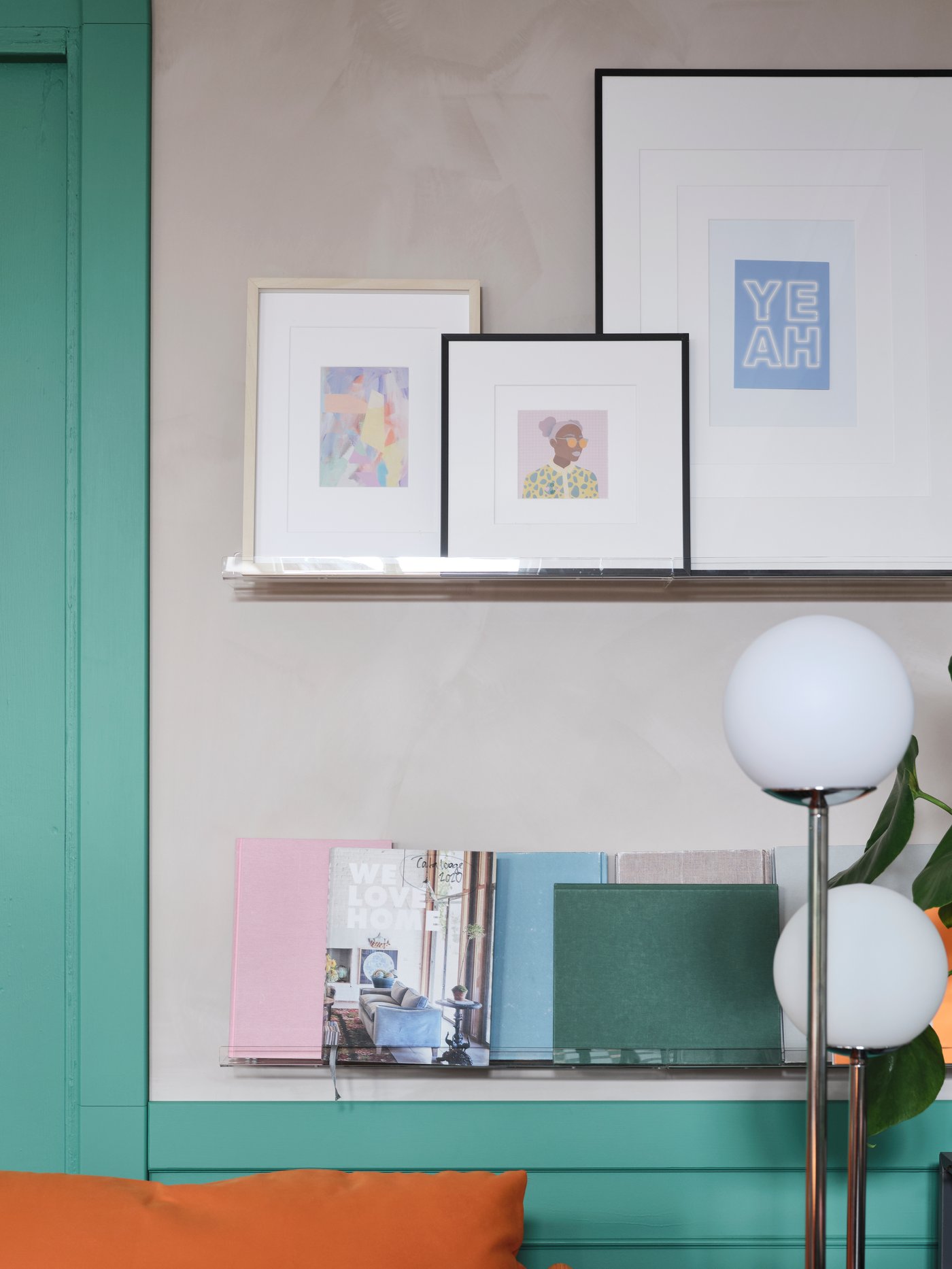 Una pared con dos estantes para cuadros transparentes, con obras de arte enmarcadas arriba y cuadernos de colores pastel abajo.