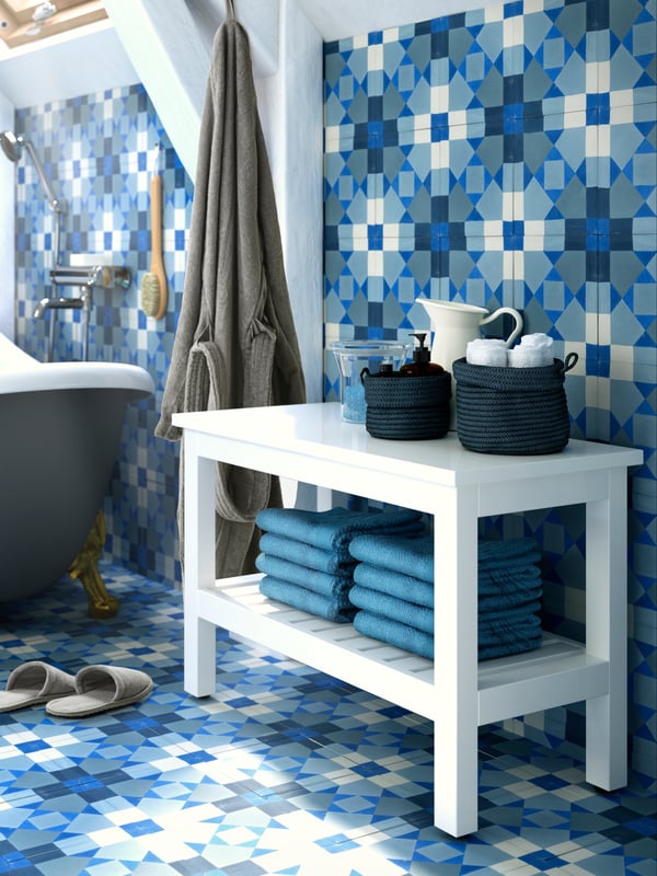 Un bagno con piastrelle azzurre geometriche. In primo piano, una panca bianca HEMNES con biancheria piegata e alcuni contenitori con oggetti da bagno. Alle travi, è appeso un'accappatoio ROCKÅN e accanto c'è una vasca da bagno. Sul pavimento, pantofole TÅSJÖN color grigio.
