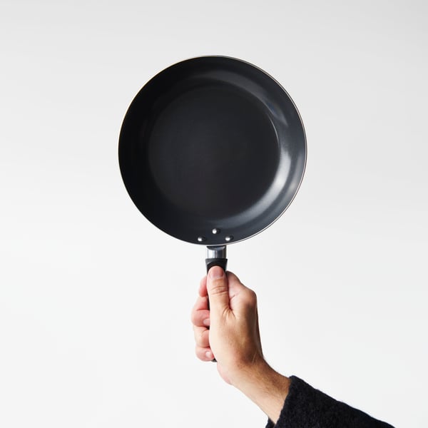 Close-up of a hand holding up a TAGGHAJ frying pan against a light-grey background.