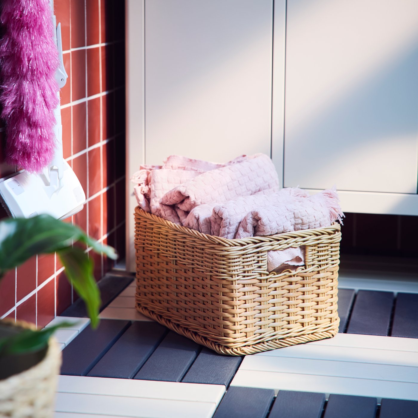A balcony with a natural coloured BEKNA basket made from plastic rattan, filled with cosy throws near a storage cabinet.
