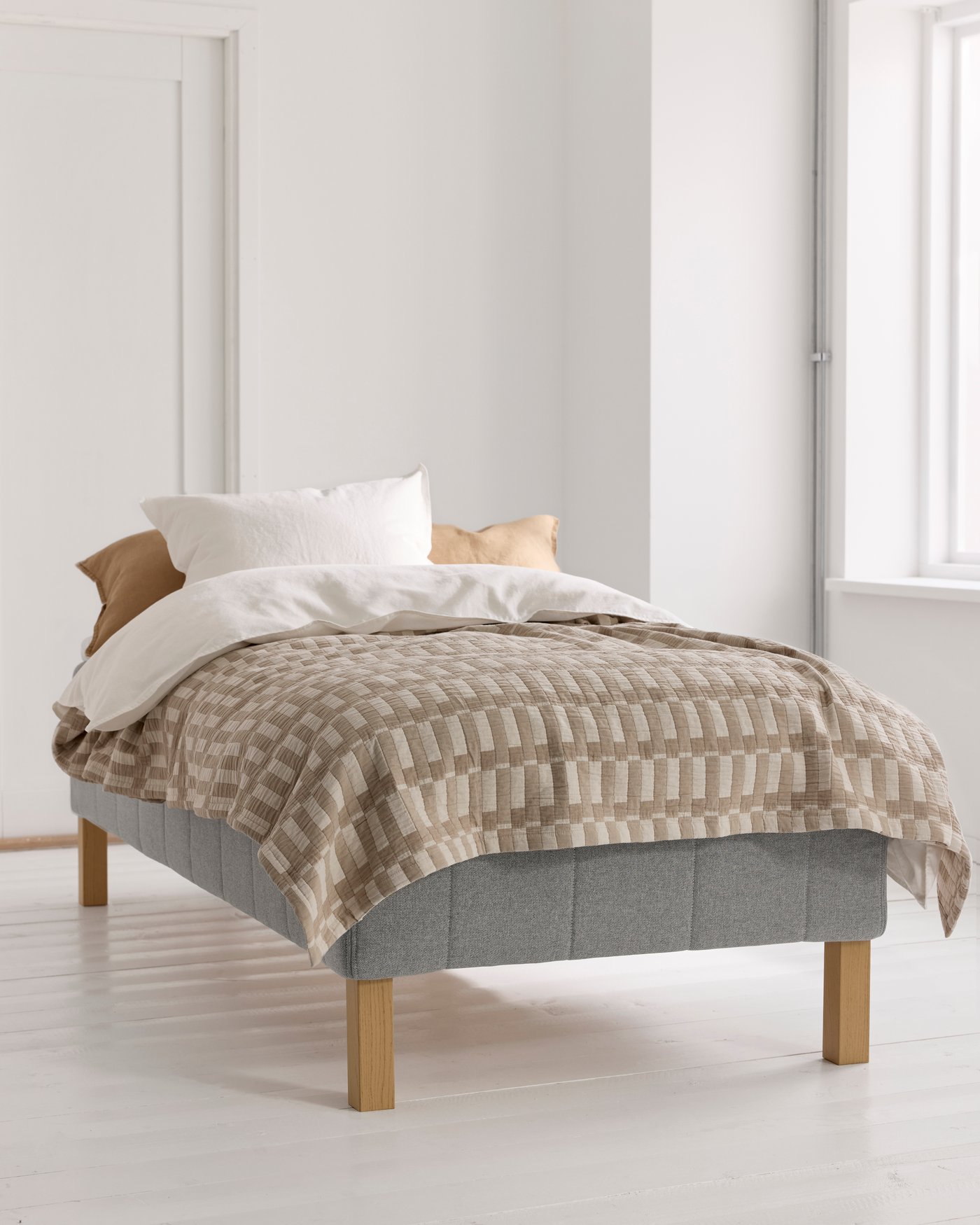 En enkelbädd med grå resårbotten och träben, bäddad med vita och beige kuddar samt ett mönstrat beige överkast, står i ett ljust rum med vita väggar och golv.
