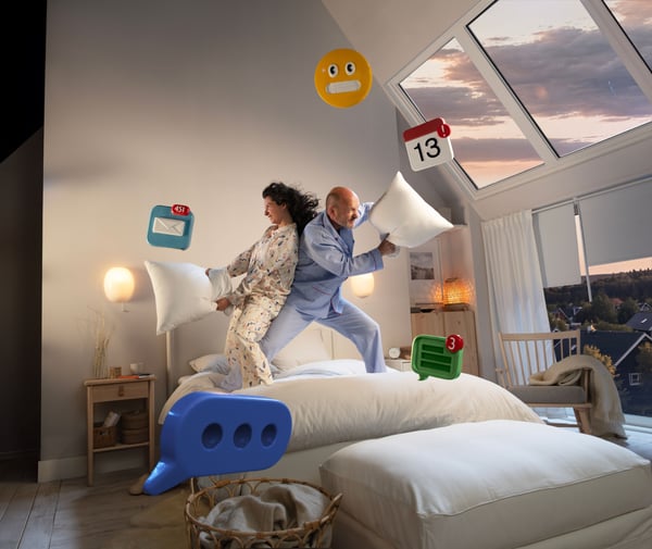 Scène surréaliste dans une chambre dont les fenêtres installées dans un plafond en pente donnent sur un ciel crépusculaire. Deux personnes sur un lit font une bataille d’oreillers, leur visage masqué par un émoji carré orange. Divers symboles 3D surdimensionnés flottent autour d’elles, comme par exemple un logo Facebook « f » bleu, une icône WhatsApp verte et une icône calendrier avec le chiffre 13. Le lit est en bataille et il y a une icône 3D bleue surdimensionnée près de lui.