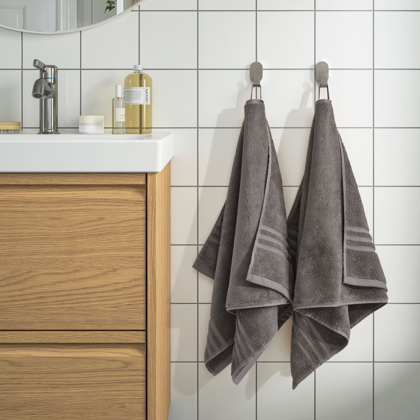 BROKGLIM Handtücher in Dunkelgrau hängen an Wandhaken neben einem Waschtisch aus Holz. Die weiche Struktur und die dezente Bordüre verleihen dem Badezimmer einen schlichten, modernen Look.