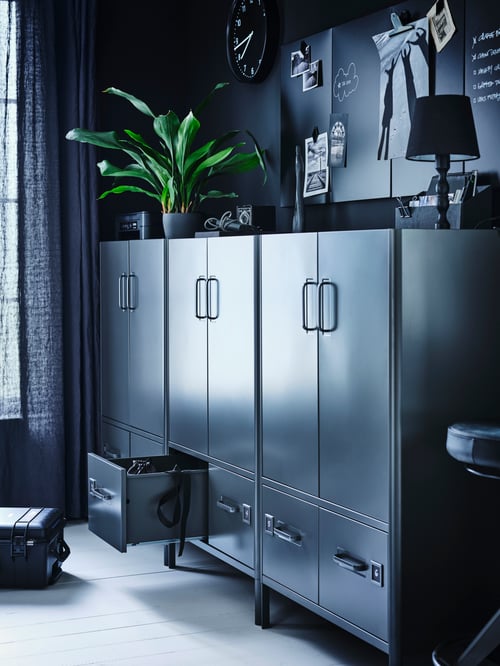 Three dark grey IDÅSEN cabinets against a wall with a plant, a lamp and stationery holder on top. One bottom drawer is open.